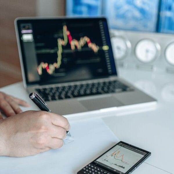 Analyst writing notes beside a laptop displaying financial market charts