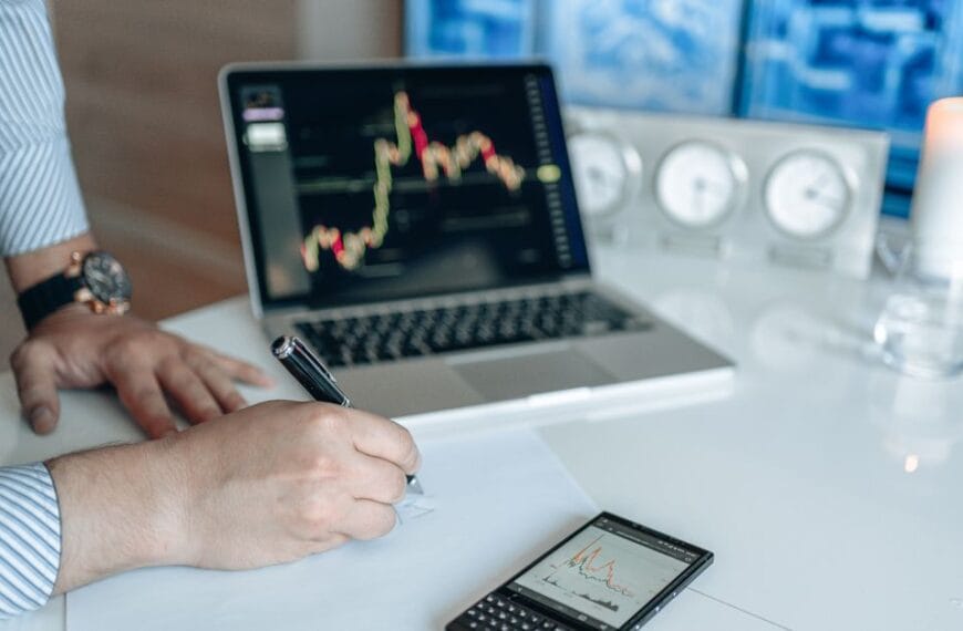 Analyst writing notes beside a laptop displaying financial market charts
