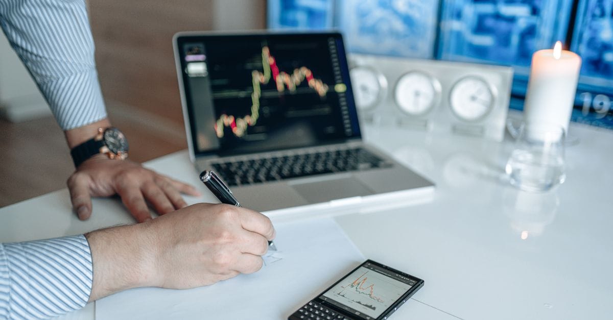 Analyst writing notes beside a laptop displaying financial market charts