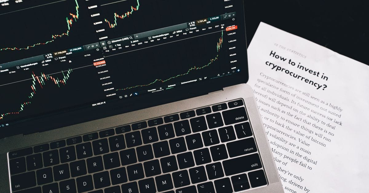 Investment research paper beside a laptop displaying financial market charts