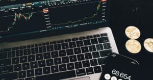 Laptop and smartphone showing cryptocurrency market data beside physical crypto tokens