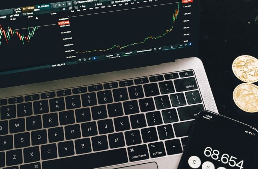 Laptop and smartphone showing cryptocurrency market data beside physical crypto tokens