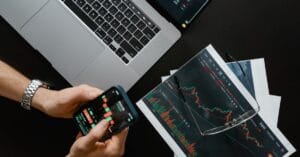 Hands using a smartphone beside a laptop displaying financial market charts