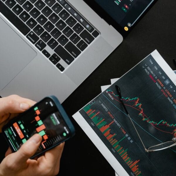 Hands using a smartphone beside a laptop displaying financial market charts