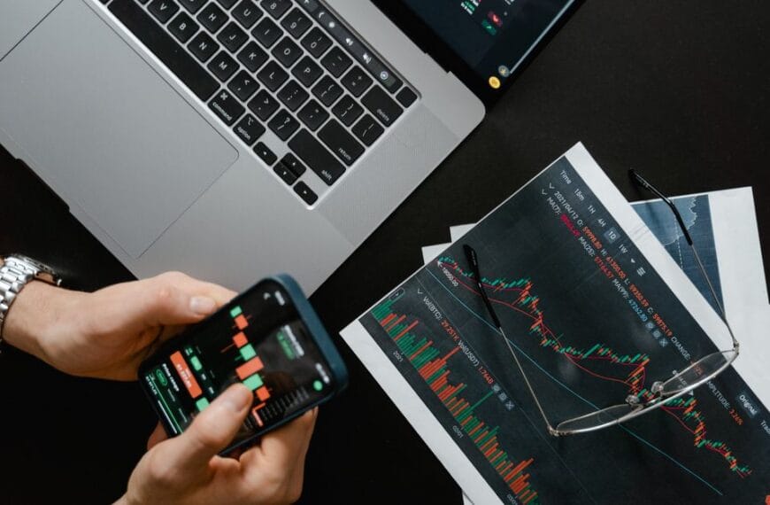 Hands using a smartphone beside a laptop displaying financial market charts