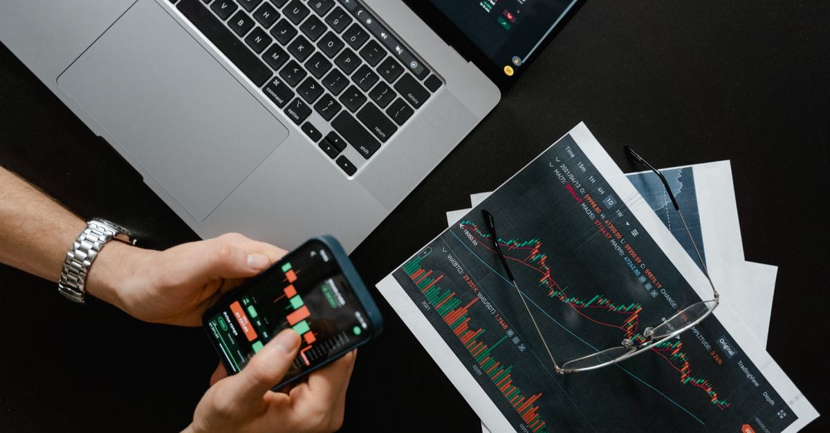 Hands using a smartphone beside a laptop displaying financial market charts